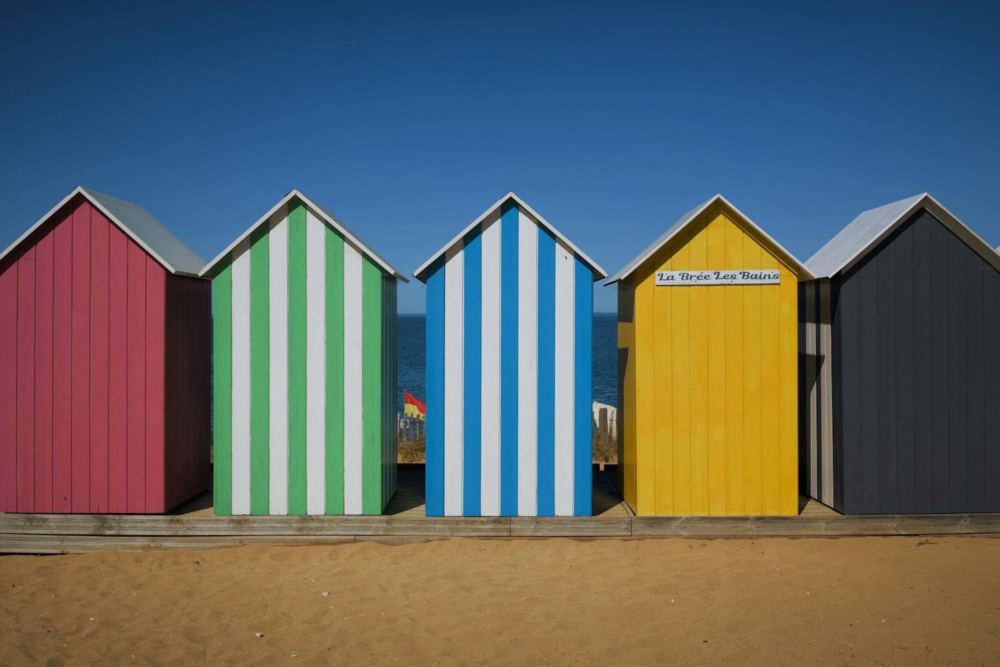 Five beach huts in a row on the sand: pink, green-striped, blue-striped, yellow and charcoal